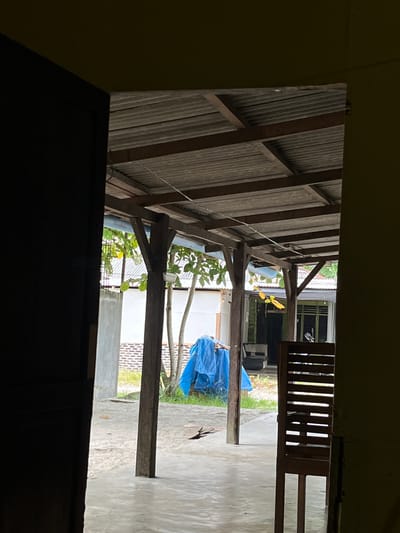 An outdoor scene is captured from a doorway, looking out onto a covered patio area with a corrugated metal roof supported by wooden posts. In the foreground, a dark doorway frames the view. The patio floor is concrete and appears to be slightly wet or reflective. Beyond the patio, there is a patch of grass and a blue tarp covering an object. Further back, a building with white walls and a brick base is visible, along with some trees with green foliage. A motorcycle is parked in front of the building. The environment suggests a residential setting in Tanjung Pura, Indonesia, possibly a home or a small business.