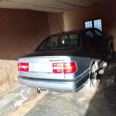 A dark grey Volkswagen Passat CL is parked in a garage. The car is facing away from the viewer, with its rear end prominently displayed. The garage has rough, textured brown walls and a white tiled ceiling. A small, barred window with a blueish tint is visible on the back wall, letting in some light. The car has its taillights illuminated, one on the left and one on the right, showing a combination of red and orange. The Volkswagen logo is visible on the trunk, along with the "Passat CL" badging. A license plate with the text "PLATEAU" and numbers is also visible. There is a tow hitch attached to the rear bumper. The floor of the garage appears to be concrete and shows signs of wetness and dirt. The lighting suggests it is daytime, with shadows cast by the car and the structure of the garage. The scene is static, with no people or activity present, implying the car is parked and likely unused. The overall impression is one of a utilitarian space for vehicle storage in a location that appears to be Nigeria, given the "PLATEAU" on the license plate which refers to a state in Nigeria.