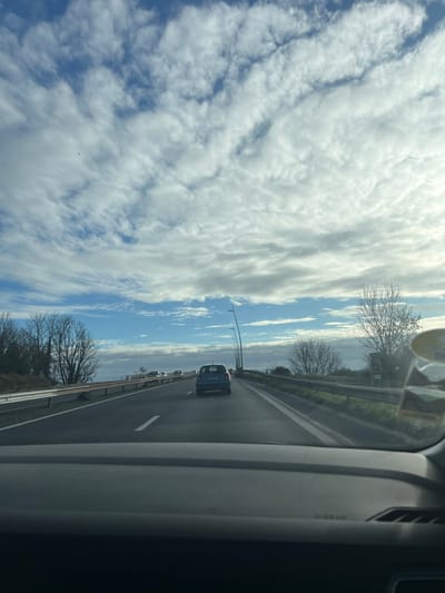The image depicts a daytime view from the interior of a vehicle on a multi-lane highway in Hérouville-Saint-Clair, France. The foreground shows the top portion of a grey vehicle dashboard. The asphalt roadway, marked with white solid and dashed lines, extends into the distance. A dark-colored hatchback, possibly a Renault Clio, is positioned directly ahead in the rightmost lane, with additional vehicles visible further down the road. Metal guardrails line both sides of the highway. The left roadside features a sloped embankment with bare deciduous trees and some evergreen vegetation. The right roadside exhibits bare deciduous trees, a series of streetlights with curved black poles, and a partially visible roadside sign. The sky above is partly cloudy, displaying patches of blue sky interspersed with widespread white and grey cloud formations. No individuals are visible in the scene.