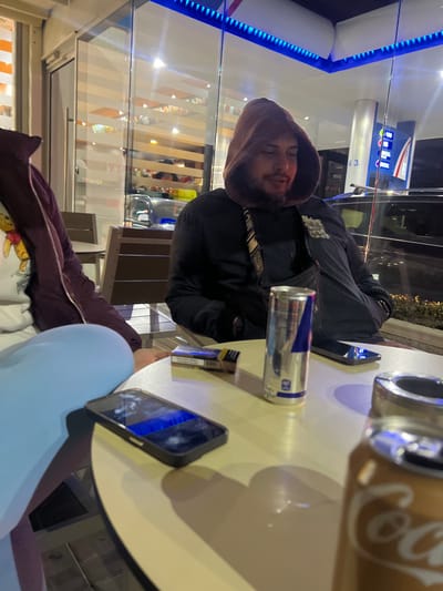 The image depicts a nighttime scene at an outdoor seating area, identified as being in Piperkov Chiflik, Bulgaria, likely associated with a gas station or convenience store.

In the center, a male individual is seated, looking slightly downward and to his right. He wears a dark hooded jacket with the hood up, covering his head, and a dark garment underneath with a patterned strap visible on his chest. He has facial hair. To his left, partially visible, is another individual wearing a maroon jacket over a white long-sleeved shirt featuring a Winnie the Pooh graphic. Light blue pants or leggings are also visible from this person.

On the light-colored round table in the foreground, several objects are present: a partially consumed silver and blue Red Bull energy drink can, a pack of cigarettes with blue and white branding, a metallic ashtray, and two smartphones. One black smartphone in the left foreground has its screen illuminated, displaying a blue and white image. Another black smartphone is further back on the table, with its screen off. A gold-colored Coca-Cola can is partially visible and out of focus in the extreme lower right foreground.

The background reveals a commercial setting. Behind the central male, gas station fuel pumps are visible, one marked with the number "3" and indicating "100," alongside a black vehicle. Bright overhead lighting illuminates the area. Behind the individual on the left, a glass storefront with horizontal opaque strips and interior lighting suggests a retail or food establishment. Blue LED strip lighting runs along the underside of the overhead structure above the seating area. Wooden slat-back chairs are partially visible in the environment.