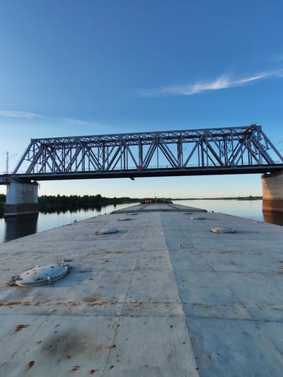 The image shows a large cargo barge on a river, moving towards a railway bridge. The barge has a flat, gray deck with several circular hatches visible. The bridge, made of metal trusses, spans the river and appears to be a significant structure. The surrounding environment consists of a wide river, a tree-lined bank on the left, and a clear blue sky with a wispy cloud formation. The lighting suggests it is either early morning or late evening, given the low angle of the sun casting shadows and illuminating the bridge. The barge is empty and appears to be in transit. There are no people visible on the barge or in the immediate vicinity of the river. The location is identified as Tarkanovo, Russia.