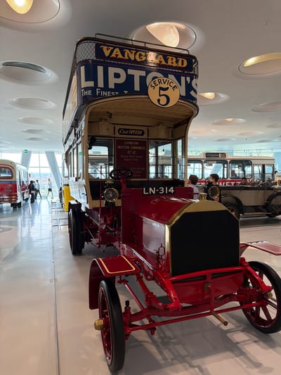 The image shows a red vintage double-decker bus, likely from the early 20th century, on display in a modern museum. The bus has "VANGUARD LIPTON'S SERVICE 5" written on its upper deck and "LN-314" on its side. Behind it, another vintage bus with "56" on its front is visible, as is a red double-decker bus on the left. The museum features a high, modern ceiling with circular light fixtures. The floor is polished and reflective. In the background, people are visible observing the exhibits. The context of the image indicates that it was taken in Stuttgart, Germany, at the Mercedes-Benz Museum, which features a wide array of historical vehicles.
