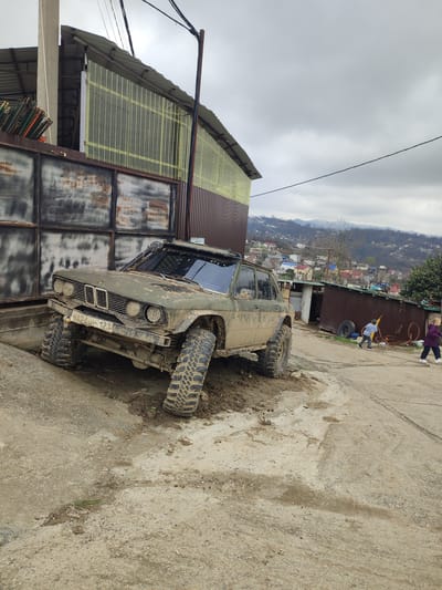 This image depicts a scene in the city of Bestuzhevskoye, Russia, under an overcast sky. The primary subject is a heavily mud-covered, olive-green custom off-road vehicle, appearing to be a modified sedan, possibly a BMW, with a Russian license plate "У24ЧЮ 123" on its front bumper. The vehicle is positioned on an incline, with its front left wheel significantly elevated and its front right wheel sunk into mud. It features large, aggressive off-road tires, a visible winch on the front bumper, a snorkel on the passenger side, and a roof rack.

To the left of the vehicle, a tall concrete pole stands adjacent to a rusty metal gate or wall. Behind this, a structure with a corrugated metal roof and translucent yellowish corrugated plastic siding is visible, with stacked metal pipes to the far left. The ground in the foreground and around the vehicle is a mix of dirt and mud, showing tire tracks and small debris.

To the right, a dirt road slopes gently downwards. Two individuals, appearing to be children, are present on this road. One child in a light blue shirt and dark pants is running rightward, holding a yellow object. Another child in a purple jacket and dark pants walks rightward. In the mid-ground, several corrugated metal structures, including what appears to be a shed or small building, are visible. An old tire and various discarded items are scattered near these structures. The background reveals a mountainous or hilly landscape dotted with numerous residential buildings under the grey sky. A utility wire stretches across the upper mid-ground.