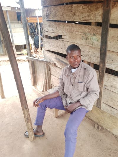 In Agwala, Nigeria, a man sits on a wooden bench in front of a weathered wooden structure. He is wearing a light brown jacket over a white undershirt and purple pants. His feet are clad in black flip-flops. The wooden structure behind him has horizontal planks with writing scrawled on them. To his left, a thick wooden pole stands upright. The ground is sandy earth. In the background, a glimpse of other structures and some people are visible through an opening. The scene appears to be an outdoor marketplace or community area.