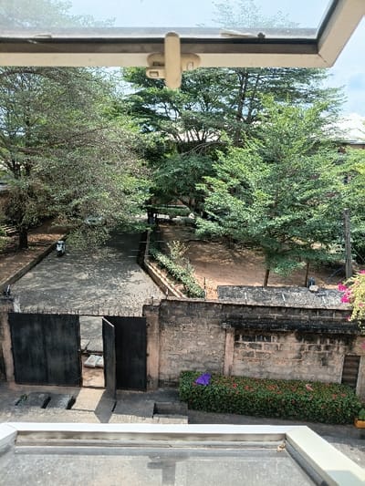 The scene depicts an outdoor environment in Oba, Nigeria, viewed from an elevated perspective, likely a window. In the immediate foreground, a light-colored window frame with a central cream-colored latch mechanism is visible. Below this frame, a grey paved surface features two rectangular metal storm drain grates. A low concrete step descends to a landscaped garden bed containing dense green foliage and small clusters of red flowers. A purple fabric or object is partially discernible within the foliage on the left side of this bed.

Behind the garden bed, a weathered concrete block wall, topped with barbed wire or razor wire, extends across the midground. Integrated into this wall is a large, dark-colored metal gate with two panels; the right panel is partially ajar, providing a glimpse into the area beyond. A white scooter is parked on a paved driveway immediately behind the gate, facing away from the viewer. To the right of the paved driveway, an unpaved section of reddish-brown earth with sparse vegetation is visible. The background is dominated by numerous trees displaying various shades of green foliage. Through and above the trees, sections of buildings with light-colored, possibly pitched, roofs are visible, with one building exhibiting reddish-brown exterior walls. The sky above is clear and light blue. No individuals are present in the depicted scene.