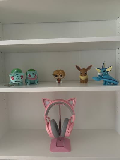 The media shows a collection of Pokémon figures and a pair of pink cat-ear headphones arranged on white shelves. On the top shelf, a blue Vaporeon figure is positioned to the left, and a brown Eevee figure is to the right. The middle shelf displays a blond-haired character figurine, possibly from an anime or video game. Below that, two Bulbasaur figures are visible. To the right of the Bulbasaur figures, a pink headset with cat ears is displayed on a stand. The background consists of plain white shelves, suggesting an indoor setting, likely a room or display area. There is no indication of time of day or weather. The scene is static, with no activity taking place. There is no visible text in the image.