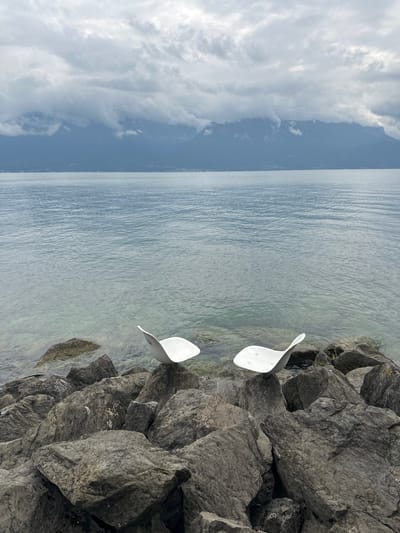 The image displays a static outdoor scene on the rocky shoreline of Lake Geneva in Vevey, Switzerland. In the foreground, a natural breakwater consists of numerous dark grey, angular boulders. Resting upon these rocks are two distinctive, white, smooth, concave objects, abstractly shaped like seats. The object on the left prominently displays the word 'VEVEY' on its visible surface. The object on the right is positioned slightly lower and further from the viewer. Beyond the rocky shore, the calm, light blue-green water of Lake Geneva extends horizontally, with the shallow areas near the rocks appearing transparent. In the distant background, a range of dark mountains is visible, partially obscured by dense, low-hanging grey clouds that fill the sky overhead. No individuals are present in the scene.
