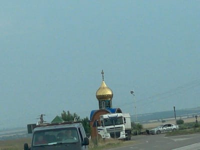 The media file depicts an outdoor scene with a clear, overcast sky suggesting a daytime setting. In the foreground, a dark-colored vehicle, possibly a van or SUV, is partially visible on the left, with people visible inside. Further to the right, a white truck is parked next to a small, domed structure that resembles a chapel or a roadside shrine. The dome is golden, and the structure has blue trim. Behind the truck and chapel, a silver car with a camouflage-like pattern is parked on the right side of the road. The background shows a rural landscape with fields and trees, indicative of a setting outside a major urban area. The overall impression is of vehicles stopped near a religious landmark along a road, possibly in a rural part of Congaz, Moldova, given the location context. There is no visible text in the media file.