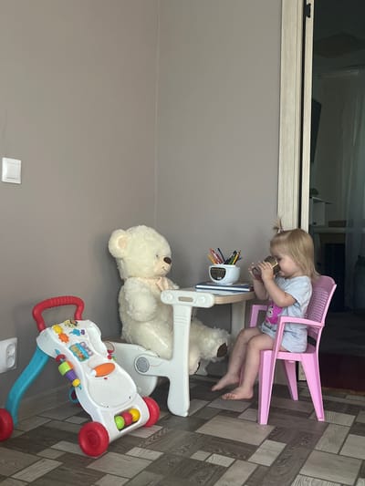 A young child with blonde hair pulled into a ponytail sits on an upside-down pink plastic chair, leaning forward to drink from a small cup. The child wears a light blue t-shirt with a pink design on the front. A toy desk with colored pencils in a dispenser is positioned in front of the child. To the right of the desk, a large, fluffy white teddy bear is lying down. In the foreground, a white and colorful baby walker with red wheels and handles is visible. The scene appears to be indoors, with a grey wall on the left and a tiled floor on the right. A light fixture is visible on the grey wall. The image is oriented vertically.
