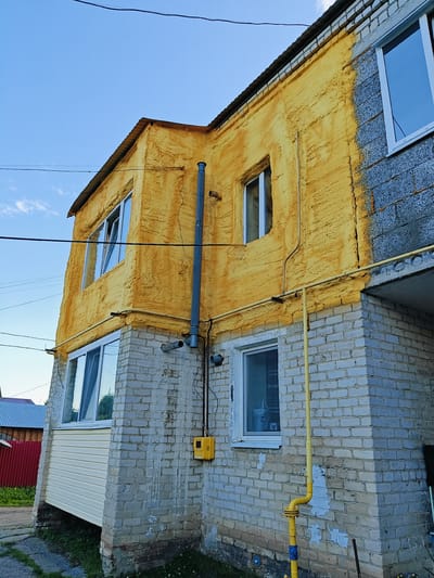 An exterior view of a multi-story building is captured from a low angle, with a clear blue sky above. The building features a section of wall covered in a rough, textured yellow insulation, contrasting with exposed grey brickwork on other parts. A metal chimney pipe runs vertically along the yellow section of the wall, attached to a yellow electrical box. Windows are visible on both levels. A yellow gas pipe runs horizontally across the building and then vertically down the brick facade, connected to a meter. The ground level shows a base of grey brick, with grass and some paving stones. In the background, a red fence and part of another building with a corrugated metal roof are visible. The scene appears to be in a residential area.