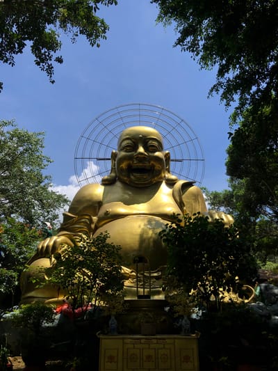The image shows a large, golden statue of the Laughing Buddha, framed by lush green trees and a clear blue sky. The statue is seated and appears to be made of polished metal, reflecting the sunlight. A circular metal structure, possibly a halo or part of an artistic installation, is visible behind the Buddha's head. In the lower part of the image, the base of the statue is surrounded by rocks, foliage, and what appear to be offerings or decorative elements. The scene is set in a garden or temple complex, with the overall impression being one of serenity and grandeur. The location is identified as Nam Ban city, Vietnam.