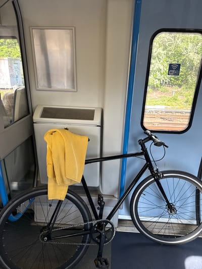 A black bicycle is secured inside a train carriage in Welwyn Hatfield, United Kingdom. The bicycle is held vertically against the wall. A yellow striped shirt is draped over part of the bike and a storage compartment. The train carriage features a window with a WiFi sticker and a view of trees. A silver panel is on the wall, next to the storage compartment.