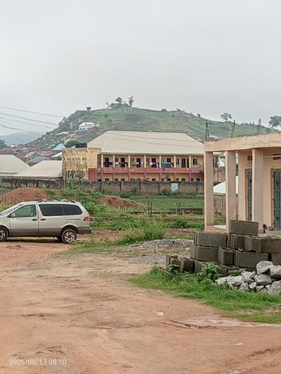 The image depicts a scene in Ibo, Nigeria, characterized by a somewhat overcast sky. In the foreground, a dirt road with visible potholes leads towards a construction site. To the right, a pile of grey cinder blocks and loose stones sits on a patch of grass, indicating ongoing building activity. Further into the scene, a silver minivan is parked on the dirt road, facing left. Beyond the minivan, a large, multi-story building with a light-colored roof and yellow and pink accents dominates the midground. This structure appears to be a school or residential complex, with visible windows and balconies. The background features a hilly landscape dotted with smaller buildings and trees, suggesting a semi-urban or semi-rural environment. The overall impression is one of a developing area with visible construction and a backdrop of natural topography.