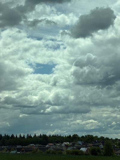 The image shows a daytime view of Юзуфоўскі сельскі Савет, Belarus. The sky is predominantly covered with clouds, ranging from light white to dark gray, with a small patch of blue visible. On the right side of the image, there is a line of trees and houses, indicating a residential area. The grass is green and lush. The image is oriented sideways.