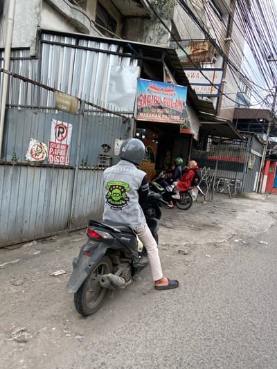 A man wearing a motorcycle helmet, a grey denim jacket with a green patch that says "Zombie Is Life," and beige pants sits on a black motorcycle. He is facing the left and looking down. The motorcycle is facing the right. The man is standing on the ground with his right foot on the motorcycle's foot peg and his left foot on the ground. The man is in front of a building with a sign that reads "Rumah Makan Saribu Bola" in red and white letters. The building is located at -6.965, 107.577 in Margahayu, Indonesia. There is another motorcycle to the right of the man, partially obscured by a car. There is a sign on the building to the left that reads "Dilarang Parkir Disini" in black and white. There are also some power lines strung overhead. The street is paved and dirty. There is a concrete curb on the right side of the road. The sky is overcast and the air is hazy. The man is parked in front of a restaurant.
