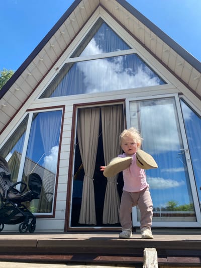 A young child stands on a wooden deck next to a building with prominent windows, under a clear blue sky. The child, a toddler with blonde hair, is wearing a pink long-sleeved shirt, tan pants, and light-colored shoes. They are holding two large, light-brown, boat-shaped objects, one on each side of their head, resembling oversized clogs or wooden shoes, possibly in playful imitation of wings. The child looks directly at the camera with a calm, curious expression.
The background features the modern architecture of a building with an A-frame or gabled roof. Its large windows, framed in white with a darker brown trim, reflect the bright blue sky and white clouds, suggesting a sunny day. Light-colored panel siding covers parts of the building. A string of small, white outdoor lights runs along the underside of the roof eaves. On the wooden deck, partially visible below the child, is a black and beige stroller or pushchair. The scene takes place outdoors, possibly at a holiday home or modern cottage in Suzdal, Russia. There is no visible text in the image.