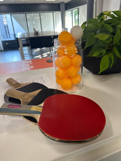 The image depicts a close-up view of a white tabletop with several ping pong paddles and a jar filled with orange ping pong balls. A green leafy plant in a dark pot is visible in the background. The environment appears to be an office or indoor recreational space with modern office furniture and large windows suggesting a well-lit room. The text on the paddles and some papers on the table indicate branding and potentially documents related to the setting. The overall scene is static and does not depict any activity or individuals. The location is not identifiable as Staphorst, Netherlands from the visual cues.