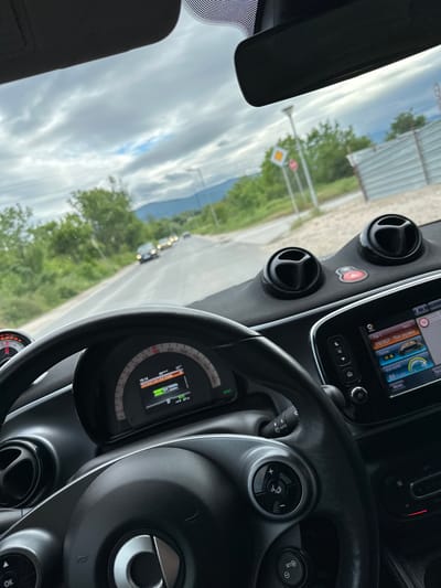 Here is a detailed description of the image:
**Scene:**
The photograph is taken from inside the cockpit of a car, with the dashboard and steering wheel dominating the foreground. Outside, the vehicle appears to be in transit on a road.
**Objects:**
- Car: The dashboard, steering wheel, and instrument panel are prominent, indicating a Smart car.
- Road: A road sign can be seen through the windshield.
- Landscape: Green foliage, possibly trees and grass, lines the road. In the distance, mountains are visible.
- Traffic: Other vehicles are present on the road, as indicated by distant headlights.
- Navigation system: The car has a navigation system running.
**Details:**
- Road sign visible shows a yellow and white diamond.
- The car is traveling on a road lined with green foliage, possibly trees and grass.
- The photograph is taken during daylight hours under a partly cloudy sky.
-The dashboard is neat and has air vents and a start button.
**Location**
The description does not provide explicit clues that point to a specific location within Plovdiv, Bulgaria, or the city itself.