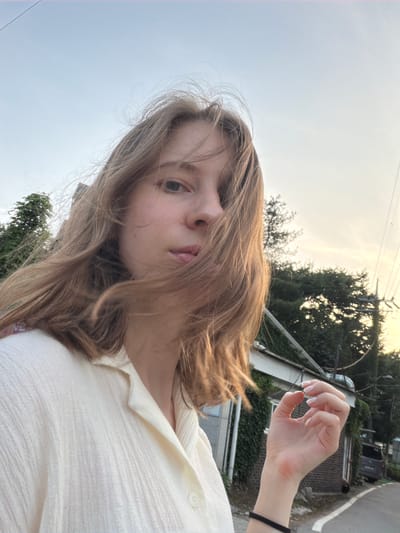 A young Caucasian woman with light brown hair is captured in a selfie outdoors during daylight. She is wearing a cream-colored shirt. Her hair is blowing across her face, obscuring part of her features. Her right hand is raised, with her thumb and index finger forming a circle. The background features a building covered in ivy, trees, power lines, and a street with a parked vehicle. The setting appears to be a rural or suburban area in Pocheon-si, South Korea. The sky is a pale blue with a hint of yellow suggesting late afternoon or early evening light.