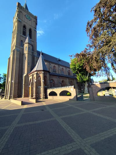 This media file is a low-angle, full shot of a large brick church with a tall clock tower. The church is constructed of red-brown bricks and has a dark-colored roof. The clock tower is prominent, featuring a clock face with a green dial and Roman numerals, and several arched openings.

The scene is outdoors, set against a clear, bright blue sky with a few wispy clouds, suggesting it is daytime and the weather is fair. Sunlight casts long shadows across the paved courtyard in front of the church, indicating it is likely morning or late afternoon. The courtyard is paved with brickwork arranged in a patterned grid.

To the right of the church, a large tree with reddish-brown leaves partially obscures the view of a brick wall with an arched opening. Beyond this wall, gravestones are visible, suggesting the church is part of a cemetery or has grounds that include one. A metal gate with an ornate cross design is open, leading into the cemetery area.

The architecture of the church, with its pointed arches and gothic-inspired windows, suggests it is a historical religious building. The overall mood is serene and peaceful, with the bright sunlight and clear sky creating a pleasant atmosphere. There is no visible text in the image. The location is identified as Schaijk, Netherlands.