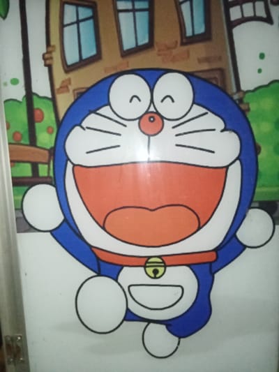 A vertically oriented photograph displays a two-dimensional cartoon illustration of the character Doraemon. Doraemon, rendered in blue and white, occupies the central foreground with its arms raised and legs positioned as if jumping. The character exhibits a wide-open red mouth, a red nose, white eyes with small black curved lines above, and black whiskers. A red collar with a yellow bell is visible around its neck, and a white utility pocket is on its lower torso. The background features a cartoon-style outdoor urban environment, including sections of a light-brown brick building with rectangular windows, green foliage with red circular elements (possibly fruit), and a brown bench. A vertical streak of light glare is present across the central portion of the image. This depiction is observed on a surface within the city of Ciputri, Indonesia.