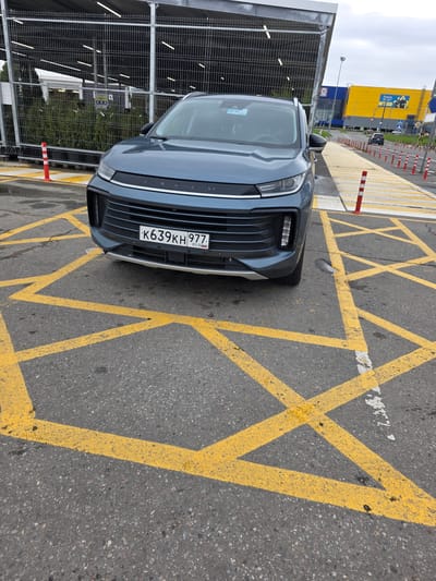 A gray SUV is parked in a parking lot with yellow painted markings. The SUV has a license plate that reads "K639KH 977". In the background, there is a large building with yellow and blue sections, likely a retail store. There are also metal fences and trees visible. The sky is overcast. The scene appears to be in an outdoor commercial area in Sholokhovo, Russia.