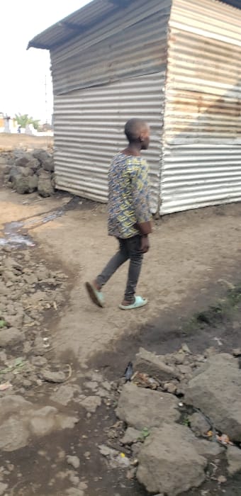 The image depicts a daytime scene in Buzove, Democratic Republic of the Congo. A young individual with short hair, wearing a multi-colored patterned long-sleeved shirt and dark trousers, is walking right-to-left across the frame on a dirt path. The person is wearing light blue open-toed sandals. The ground is uneven, composed of packed dirt, loose stones, and areas with standing water or damp soil. To the left of the path, a collection of dark, irregular rocks or rubble is present. In the background, a structure constructed from corrugated metal sheets with a sloped roof is visible, partially obscured by the individual. Further in the background, out of focus, appear to be more structures and some sparse vegetation under an overcast sky.