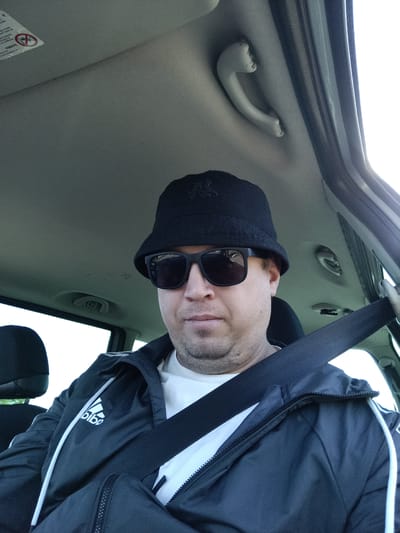 A medium shot shows a man sitting in the passenger seat of a vehicle, looking towards the camera. He is wearing a black bucket hat, black sunglasses, a white t-shirt, and a black Adidas jacket. A black seatbelt is fastened across his chest. The interior of the car, including the ceiling with a handle and a sun visor sticker, is visible. The scene appears to be in Čistá, Czechia.