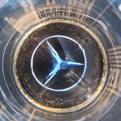 This image provides an extreme close-up, overhead view of the interior of a clear plastic bottle, likely a beverage bottle, looking down into its base. The primary subject is the embossed logo on the bottom of the bottle, which is a stylized three-pointed star within a circle, illuminated with a bright, cool blue light, making it stand out sharply against the darker interior. This logo is characteristic of the Mercedes-Benz brand.
The bottle itself is transparent, and its inner walls show a textured pattern, suggesting a common design for plastic bottles. The exterior of the bottle is out of focus, but appears to be cylindrical with some ribbed or grooved details. The lighting within the bottle is dynamic, with reflections and highlights playing across the inner surfaces, creating a somewhat abstract and visually interesting pattern. There are small water droplets visible on the inner surfaces, especially around the rim of the bottle's base, and some darker residue or sediment is present at the very bottom, surrounding the central logo.
The overall scene is a still life, focusing entirely on the details of the bottle's interior. There is no indication of time of day, weather, or specific location beyond the inferred context of Germany. No discernible text is visible on the logo or the bottle itself within the frame, though it is possible that such text might be present on other parts of the bottle not captured in this close-up view. The image captures a simple object in a visually amplified way, drawing attention to its design and the play of light.