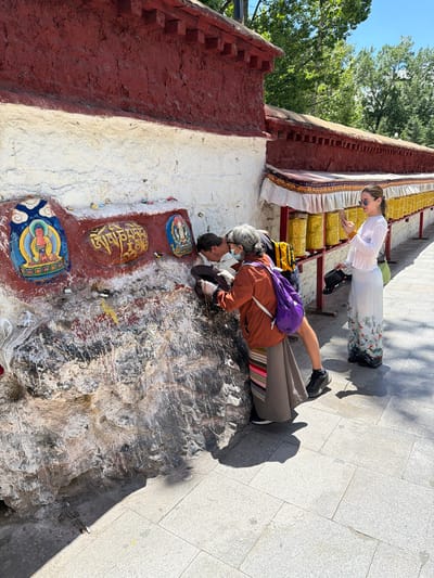 The image captures an outdoor scene on a bright, sunny day, likely at a Tibetan Buddhist site. Two individuals are present on a light grey tiled walkway next to a building or wall adorned with religious elements.
One person, appearing older with grey hair, is seen from behind, leaning forward intently towards a painted section of the wall. They are wearing a brown jacket, grey trousers with a colorful striped design at the hem, a purple backpack, and a face mask. Their hands are positioned near the base of the wall, where there are three colorful, painted or carved images. The central image clearly displays the Tibetan mantra "Om Mani Padme Hum" in golden script on a red background, flanked by two other painted figures resembling Buddhas or Bodhisattvas. The person's posture suggests a moment of contemplation, prayer, or veneration.
Standing slightly behind and to the right is another individual, dressed in a long-sleeved white top and a long white skirt with an elaborate floral and bird pattern at the bottom. They are wearing sunglasses and are positioned near a row of golden prayer wheels that line the wall. Their left hand appears to be resting near the prayer wheels, while they hold a black hat in their right hand.
The scene is illuminated by strong sunlight, casting distinct shadows on the ground. In the background, vibrant green trees are visible against a clear blue sky, indicating an outdoor, natural setting. The overall atmosphere suggests a moment of cultural or religious observance.