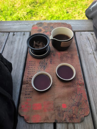 This media file shows a tea set arranged on a wooden surface, likely outdoors. The tea set includes a creamer or small pitcher containing a dark liquid, a lid, and two small cups also filled with the same dark liquid, likely tea. The objects are placed on a rustic, brown mat with an intricate design that appears to be a tree or branches, along with some red stamps or seals and what looks like handwritten calligraphy in Chinese characters.

The wooden surface is made of planks, suggesting an outdoor setting like a deck or a picnic table. To the left of the wooden surface, blurred green foliage is visible, indicating a natural environment, possibly a garden or park. The lighting suggests it is daytime, and the overall scene evokes a sense of calm and relaxation associated with enjoying tea. The presence of the tea set and its arrangement implies a tea ceremony or a casual tea break.

The visible text on the mat is in Chinese characters. The primary elements are the tea set, the decorative mat, and the wooden surface, all contributing to a peaceful and cultural aesthetic.