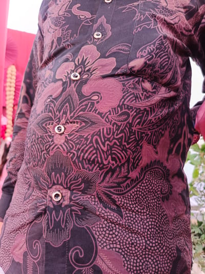 The image shows a close-up view of a person wearing a dark colored batik shirt with a floral and dragon pattern in shades of maroon and black. The shirt has several gold-colored buttons down the front. The background is blurred, with hints of pink fabric and greenery. The scene appears to be outdoors. The location in the city of Meuliek, Indonesia is not discernible from the visual information provided in the image.