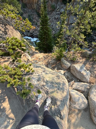 The image shows a person's legs and feet standing on a rocky cliff overlooking a waterfall. The person is wearing dark leggings and athletic shoes. The scene is set in a natural environment with pine trees and large rocks. Sunlight casts shadows, suggesting it is either morning or late afternoon. The waterfall cascades down a rocky slope, surrounded by dense vegetation. The rocky terrain implies a hiking trail or a viewpoint in a mountainous area, consistent with the Estes Park, United States location. The overall impression is one of an outdoor adventure and exploration in a scenic, natural landscape. There is no visible text in the image.
