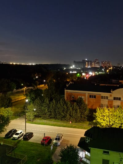 The image depicts a nighttime scene in the city of Ерино, Russia, viewed from an elevated perspective. In the foreground, a dark green roof of a building with a dormer window is visible. To the left of this building, a street is illuminated by streetlights, with several parked cars. These include two black sedans, a white sedan, a dark-colored SUV, a red sedan, and a silver SUV. Across the street, a beige building with orange brick accents and numerous windows is prominent, bordered by dense green trees. Beyond this, a line of trees separates the immediate area from a distant cityscape with illuminated multi-story buildings. The sky is dark, indicating late evening or night, with faint lights visible on the horizon and scattered in the distance.