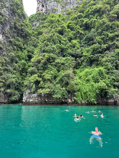 The image depicts a group of people swimming and enjoying the turquoise waters of Haad Yao, Thailand. The scene is framed by steep, jungle-covered limestone cliffs that rise dramatically from the sea. The water is clear and calm, with sunlight dappling its surface. Several individuals are visible in the water, some swimming leisurely, while others appear to be engaged in conversation or simply floating. The lush greenery of the cliffs provides a vibrant contrast to the blue of the sea and sky. The overall impression is one of a serene and idyllic tropical paradise.