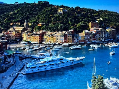This media file captures a picturesque harbor scene in Portofino, Italy, under a clear blue sky, suggesting a sunny day. The image is dominated by a large, elegant white yacht prominently positioned in the foreground, its sleek lines and multiple decks indicative of luxury. Surrounding the yacht are numerous other boats, including smaller yachts and sailboats, moored in the vibrant blue water.
The background reveals a charming Italian coastal town characterized by colorful buildings with terracotta roofs lining the waterfront. These structures are nestled against a lush, green hillside densely covered with trees. Several buildings are visible higher up the hill, some appearing to be large villas or homes, suggesting a wealthy and scenic setting. A church with a distinct dome and bell tower stands out amongst the town's architecture.
Along the stone quay in the left foreground, several people are visible strolling and gathered, adding a sense of life and activity to the scene. The overall atmosphere is one of tranquility and leisure, typical of a Mediterranean resort town. No visible text is present in the image. The time of day appears to be midday, given the bright sunlight casting shadows.