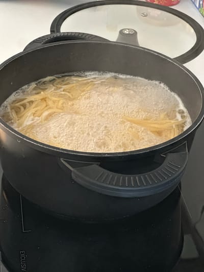 The image shows a black pot with a clear glass lid, containing spaghetti boiling in water. The pot is situated on a black stovetop, possibly an induction hob, as indicated by a faint white "EXCLUSIV" marking on the stovetop. The spaghetti appears to be al dente, with some strands submerged and others breaking the surface of the bubbling water. The scene is indoors, likely in a kitchen environment, though no specific details of the surrounding area are visible beyond the stovetop and a white countertop in the background. No people are present in the image. The overall impression is a close-up shot of pasta being cooked. There is no information in the image that can be used to determine the location in Sankt Augustin, Germany.
