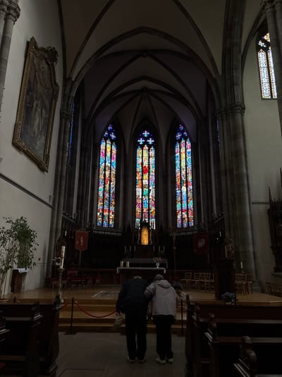 The image shows the interior of a gothic church, likely the Saint-Laurent church in Ribeauvillé, France, due to the architectural style and stained-glass windows. Two individuals, an older man and woman, are standing facing away from the camera, towards the altar. They appear to be together, with the man's arm around the woman's shoulders, and they are looking at the stained-glass windows in the background. The church features high vaulted ceilings, stone columns, and large, colorful stained-glass windows depicting religious scenes. An ornate painting hangs on the left wall. The altar area is dimly lit, with a crucifix and candles visible. Benches are arranged in rows in the foreground and midground. The overall atmosphere is serene and solemn.