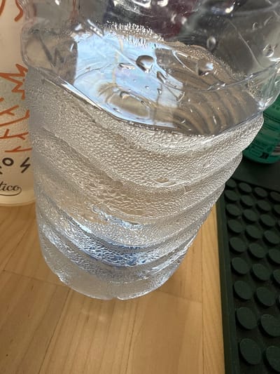 This media file is a close-up, eye-level shot of a clear plastic water bottle. The bottle is covered in condensation, with many small water droplets clinging to its surface. The bottle has a ribbed texture, and the condensation is particularly noticeable in the grooves. The bottle is lying on its side on a light wooden surface, likely a floor or table. In the background, a dark, textured object with a repeating pattern of circular indentations is visible, possibly a mat or a piece of equipment. A portion of a green item, possibly another bottle or container, can be seen in the upper left corner. The lighting appears to be natural and diffused, suggesting it could be daytime, but there are no strong shadows to indicate a specific time of day. The overall scene is simple and focused on the detail of the condensation on the water bottle, creating a sense of refreshment or a cool beverage. There is some faint, unreadable text on a white surface beneath the bottle, and a partially visible word "tico" is discernible. No people are present in the image. The location is Almada, Portugal.