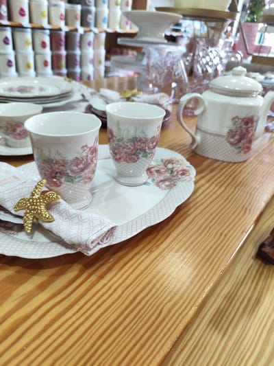 The scene depicts a tabletop display of fine china tea service, likely for sale, set against a backdrop of stacked merchandise. The primary objects include floral-patterned teacups, saucers, plates, a teapot, and a decorative gold starfish napkin ring. The items rest on a light-colored wood table with visible grain. In the background, shelves are filled with rows of uniform containers, possibly canisters or mugs, with varying color-coded labels. The setting appears to be an upscale home goods store or boutique. The image likely originates from Ponta Delgada, Portugal, given the image request context.