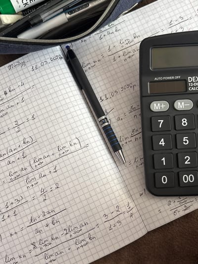 The image shows a close-up, overhead view of a notebook with mathematical equations and calculations written on its grid paper. A black pen rests on the notebook, and a black calculator is positioned to the right of the paper. The notebook also contains a handwritten date, "11.03.2020p.". Some of the equations involve limits and fractions. The background appears to be a textured brown surface. The image is well-lit, with the focus on the notebook and calculator. There is no indication of the city of Jrarat, Armenia, or any people present in the image.