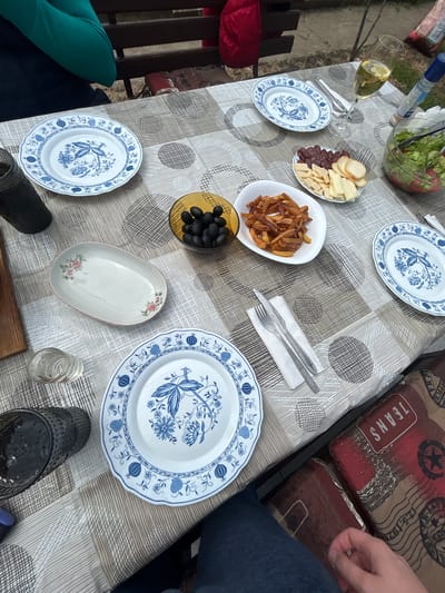 The image depicts a partially set table outdoors in daylight. The table is covered with a patterned tablecloth and various food items are arranged on it, including a bowl of salad, plates with assorted appetizers (cheese, meats, french fries, olives), and a glass of white wine. Silverware is placed beside the food. Two individuals are partially visible; one wearing jeans and another wearing a green garment. The location is Borovan, Bulgaria.
