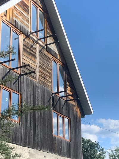 This image depicts the exterior of a multi-story building in Yasinia, Ukraine, captured from a low angle under a clear sky. The building's facade is constructed predominantly from weathered wood, featuring darker vertical planks on the lower section transitioning to lighter, varied horizontal planks and logs on the upper levels. A light grey concrete foundation is visible at the very bottom. Numerous rectangular windows with light brown frames are set into the wooden facade, reflecting a bright blue sky with scattered white clouds. Dark-colored metal brackets, consisting of horizontal bars supported by diagonal elements, are affixed to the wooden exterior, particularly between windows on the upper stories. The building features an angled roofline with white soffit and fascia visible along the right edge. Green foliage from a tree is partially visible in the lower-left and lower-right foreground. A thin dark line, consistent with a power line, is faintly visible against the sky on the right side. No individuals or specific activities are present.
