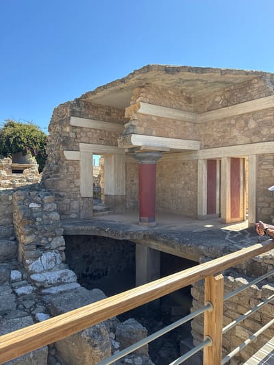 The image depicts the Knossos Palace, an archaeological site located in the city of Heraklion, Crete, Greece. The scene shows the ruins of an ancient structure, characterized by stone walls, columns, and remnants of floors and ceilings. A prominent red and dark gray column stands in the foreground, supporting a section of the roof. To the left, a stone staircase leads down into a dark, recessed area. In the background, a lush green tree with pink flowers is visible. The sky is clear and blue. A wooden railing with metal bars is positioned in the lower part of the image, suggesting that this area is accessible to visitors. The overall impression is one of historical preservation and archaeological interest.