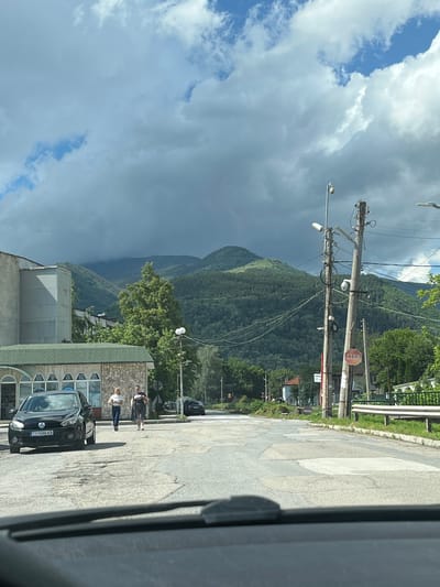 This media file captures a daytime street scene in Zlatitsa, Bulgaria, featuring a mountainous backdrop under a partly cloudy sky. The image is originally rotated 90 degrees clockwise.

In the foreground, a paved road stretches from the bottom right towards the background. On the right side of the road, a dark-colored Volkswagen car, likely a Polo, is parked, displaying the license plate `C0 5046 KB`. Next to the car, three individuals are visible. Two women are standing, one in a white top and blue jeans, and another in a darker top and skirt. A third person is partially obscured further back.

Utility poles laden with numerous wires and various devices, including what appear to be streetlights and surveillance cameras, dominate the central portion of the image. A red, octagonal "STOP" sign is affixed to one of these poles. Behind the people and car on the right, a building with a green-tiled roof and a stone-textured facade features arched windows or display cases. Another building with a flat, light-colored roof is visible to the left.

The background is characterized by lush green mountains, densely covered in trees, suggesting a hilly or mountainous terrain. The sky above is a mix of bright white cumulus clouds and darker, grayish clouds, with patches of blue sky peeking through. No specific event or intense activity is occurring; the scene depicts a static moment of daily life in a town setting nestled within a natural landscape.