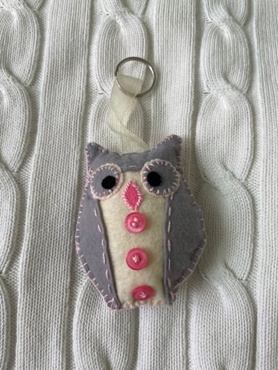 A handmade felt owl keychain rests on a white knitted blanket with a cable-knit pattern. The owl is made of gray felt with pink stitching along the edges. It has a cream-colored belly with three pink buttons and a pink felt beak. The eyes are black with gray felt outlines. A silver keychain ring is attached to the owl with a white ribbon. The background is the textured surface of the white knit blanket.