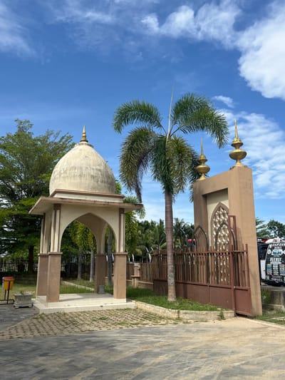 The image depicts an outdoor scene with architectural elements and vegetation under a blue sky with scattered clouds. In the foreground, a paved walkway leads towards a small pavilion with a domed roof, supported by arches. Adjacent to the pavilion is a taller, tan-colored structure with decorative arched openings and finials on top. A tall palm tree stands between these two structures. A brown metal fence runs along the right side of the scene, partially obscuring a blue van with text on its side. Lush green trees and palm trees are visible in the background, suggesting a tropical or subtropical environment. The scene appears to be a public space or an entrance to a complex.