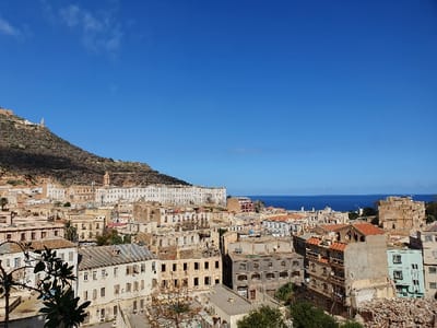 The image shows a panoramic view of the city of Oran, Algeria, taken from a hillside overlooking the Mediterranean Sea. The coordinates provided place the location near the historic center of Oran, likely the Casbah or a nearby area. The image captures a dense urban fabric, with buildings of varying heights and ages, constructed in a mix of architectural styles. The cityscape is dominated by a large, imposing building on the hilltop, possibly a former colonial administrative building or a religious structure. The sea, with its clear blue water and a distant horizon, stretches out in the background, complementing the earthy tones of the city buildings. The cityscape features a mix of residential, commercial, and institutional buildings, exhibiting signs of both decay and regeneration. Several trees are visible within the cityscape, providing some greenery. The overall scene appears to be peaceful and quiet, with no noticeable human activity in the image.