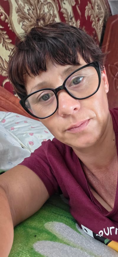 A woman with short, dark brown hair and glasses is lying on her side, looking at the camera. She is wearing a maroon t-shirt. Her left arm is resting on a pillow with a floral pattern. The background consists of a patterned wallpaper and a colorful blanket. The scene appears to be indoors, possibly in a bedroom. The image has a casual, informal feel.