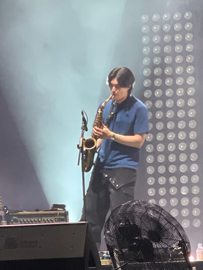 A musician plays a saxophone on a stage. He is wearing a blue short-sleeved shirt and dark pants. A microphone is positioned in front of the instrument. Behind him is a grid of illuminated lights. To his right is a large fan and stage equipment. The location is Salembaran, Indonesia.
